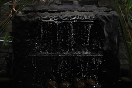 A dark water fountain with cascading water flowing down a rugged, stone-like structure. The backdrop consists of a textured stone wall with some greenery, including tall leaves and plants framing the fountain.