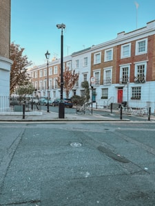 A row of elegant, terraced townhouses with a classic architectural style lines the street. The buildings are predominantly white and brick-colored, with small balconies and potted plants. A tree with autumnal foliage is visible near the sidewalk. Bicycles are parked along the street, and there is minimal traffic with a few cars parked by the curb. A streetlamp and traffic signs are present, contributing to a quaint, urban atmosphere.