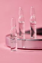 Small glass ampoules are elegantly arranged on a reflective surface with a soft pink background, creating a minimalist and clean aesthetic.