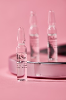Small glass ampoules are elegantly arranged on a reflective surface with a soft pink background, creating a minimalist and clean aesthetic.