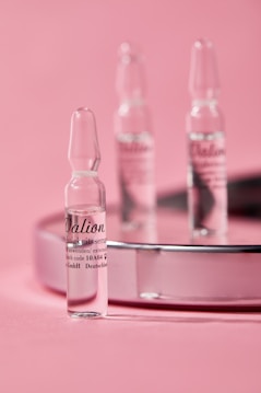 Small glass ampoules are elegantly arranged on a reflective surface with a soft pink background, creating a minimalist and clean aesthetic.