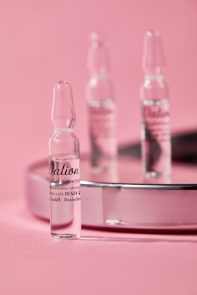 Small glass ampoules are elegantly arranged on a reflective surface with a soft pink background, creating a minimalist and clean aesthetic.