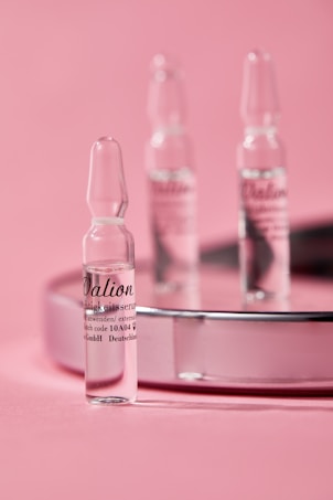 Small glass ampoules are elegantly arranged on a reflective surface with a soft pink background, creating a minimalist and clean aesthetic.