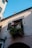 A charming European-style building facade with a small balcony. The balcony features a decorative wrought-iron railing and various potted plants. The building is a light pink color, and the balcony is shaded with a striped awning. Sunlight casts interesting shadows on the wall.
