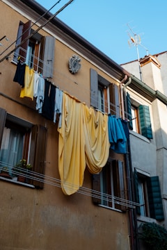 Clothes are hanging on a laundry line stretching across the facade of a building with open wooden shutters. The building is adorned with an ornamental decoration. There are a variety of garments, including shirts and large sheets in shades of yellow, blue, and black. Potted plants are visible on window sills, and the sky is clear and blue.
