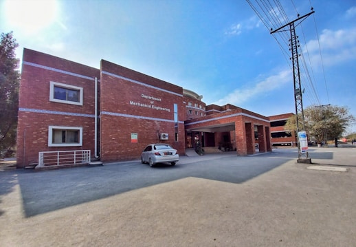 A brick building with a sign for the Department of Mechanical Engineering is situated in an open area. There is a white car parked in front of the building. The structure has multiple windows and a covered entrance. Power lines and a pole are visible on the right, with some trees in the background. The sky is clear with few clouds, indicating sunny weather.