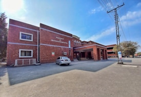 A brick building with a sign for the Department of Mechanical Engineering is situated in an open area. There is a white car parked in front of the building. The structure has multiple windows and a covered entrance. Power lines and a pole are visible on the right, with some trees in the background. The sky is clear with few clouds, indicating sunny weather.