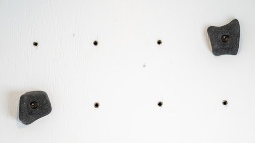 A white wall with several small black holes arranged in a grid pattern, along with two dark gray climbing holds fixed to it.