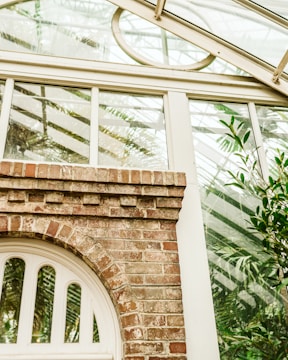 A brick archway is integrated into a glass greenhouse structure, surrounded by leafy plants. The structure features a combination of rustic brickwork and modern glass panes, allowing a view of the lush greenery inside. Sunlight creates reflections and shadows, enhancing the natural ambiance.