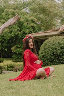 A woman in a red lace dress sits on the grass, gently cradling her belly. She is adorned with a red floral crown and surrounded by lush greenery and a rustic building in the background.
