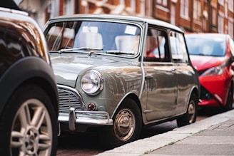 A classic Mini Cooper is parked on a city street, partially surrounded by other vehicles. Its vintage design features a compact, boxy shape with round headlights and a pastel green color. The background includes a mix of urban architecture with red brick buildings.