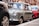 A classic Mini Cooper is parked on a city street, partially surrounded by other vehicles. Its vintage design features a compact, boxy shape with round headlights and a pastel green color. The background includes a mix of urban architecture with red brick buildings.