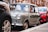 A classic Mini Cooper is parked on a city street, partially surrounded by other vehicles. Its vintage design features a compact, boxy shape with round headlights and a pastel green color. The background includes a mix of urban architecture with red brick buildings.
