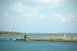 A serene coastal landscape featuring a large wind turbine situated on a distant shore. The foreground includes calm water with a series of piers and a small, abandoned building on a grassy strip. In the background, a stretch of land can be seen under a partly cloudy sky.