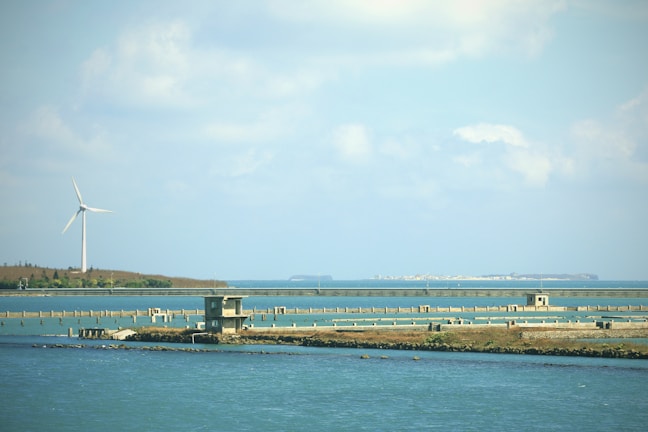 A serene coastal landscape featuring a large wind turbine situated on a distant shore. The foreground includes calm water with a series of piers and a small, abandoned building on a grassy strip. In the background, a stretch of land can be seen under a partly cloudy sky.
