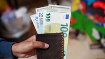 A hand is holding a black wallet from which several Euro banknotes are visible, including denominations of 20 and 100 Euros. The background is blurred, containing colors like green and red, possibly indicating a casual indoor setting.