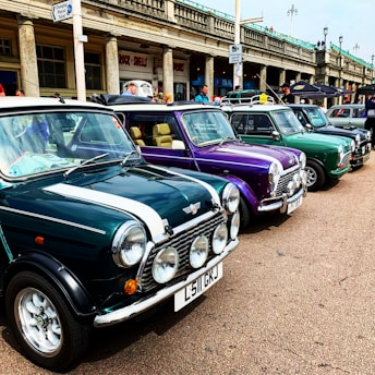 A row of vintage cars is parked along a seaside promenade. The cars, including classic Minis, are in various colors such as green and purple. The setting is a lively street scene with people walking and observing the cars. The background features historic architecture with signs and shops.