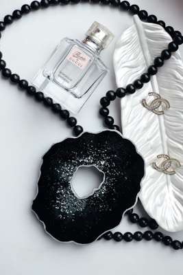 A glass bottle of Gucci Flora perfume is placed on a flat surface, surrounded by a string of black beads. An ornamental black object resembling a geode is positioned next to a white ceramic leaf dish, which holds a pair of gold earrings with rhinestones.