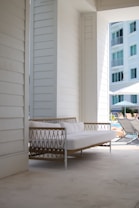 A modern outdoor patio area featuring a white, cushioned sofa with a woven design, situated under a shaded structure. The building has white paneling, and in the background, a multi-story building with balconies is visible. Patio loungers and umbrellas can be seen near a pool, suggesting a relaxing and leisurely setting.