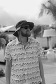 A person with a beard wears a tropical-themed shirt and dark sunglasses, accessorized with a wide-brimmed hat. The background features blurred palm trees and an outdoor setting, giving a vacation-like atmosphere.