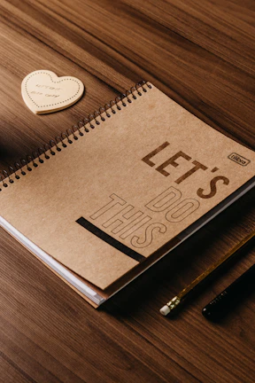 A spiral-bound notebook with a cardboard cover is placed on a wooden table. The cover has the words 'LET'S DO THIS' printed in bold. Nearby, there is a heart-shaped notepad with the words 'LITTLE MOMENTS'. A pencil and a pen are also visible on the table.