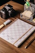 A wooden desk with a neatly arranged set of items including a 2023 planner with a checkered design, a Canon camera, an eraser, a pencil, and a pen holder containing various office supplies like pens, sticky notes, and clips.