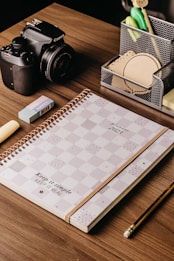 A wooden desk with a neatly arranged set of items including a 2023 planner with a checkered design, a Canon camera, an eraser, a pencil, and a pen holder containing various office supplies like pens, sticky notes, and clips.