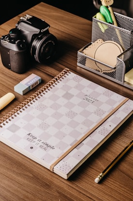 A wooden desk with a neatly arranged set of items including a 2023 planner with a checkered design, a Canon camera, an eraser, a pencil, and a pen holder containing various office supplies like pens, sticky notes, and clips.