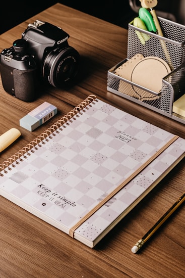 A wooden desk with a neatly arranged set of items including a 2023 planner with a checkered design, a Canon camera, an eraser, a pencil, and a pen holder containing various office supplies like pens, sticky notes, and clips.
