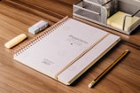A spiral-bound planner with the words 'Happiness from the Heart' lies on a wooden desk. Next to it are a yellow highlighter, a blue eraser, a wooden pencil, and a metallic stationery organizer containing sticky notes.