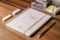 A spiral-bound planner with the words 'Happiness from the Heart' lies on a wooden desk. Next to it are a yellow highlighter, a blue eraser, a wooden pencil, and a metallic stationery organizer containing sticky notes.