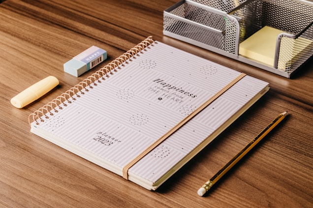 A spiral-bound planner with the words 'Happiness from the Heart' lies on a wooden desk. Next to it are a yellow highlighter, a blue eraser, a wooden pencil, and a metallic stationery organizer containing sticky notes.