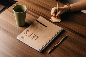 A notebook with the words 'LET'S DO THIS' on its cover is placed on a wooden table alongside a green cup, a pencil, and a hand writing on a circular notepad. The lighting is warm and emphasizes the wooden textures.