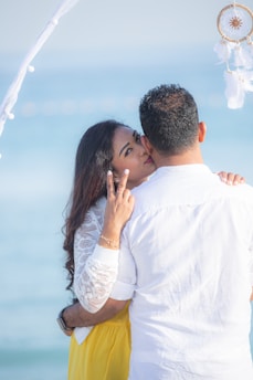 A couple is embracing by the seaside with a dreamcatcher hanging nearby. The woman is smiling and gesturing peace with her fingers. They are dressed in casual, summer attire.
