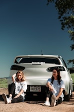 Two people are sitting on the ground in front of a white car with a visible Colorado license plate. They are wearing casual white t-shirts and black pants, with trees and open sky in the background. The setting appears to be an outdoor area with dirt and sparse vegetation.