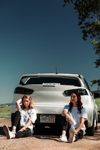 Two people are sitting on the ground in front of a white car with a visible Colorado license plate. They are wearing casual white t-shirts and black pants, with trees and open sky in the background. The setting appears to be an outdoor area with dirt and sparse vegetation.