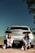 Two people are sitting on the ground in front of a white car with a visible Colorado license plate. They are wearing casual white t-shirts and black pants, with trees and open sky in the background. The setting appears to be an outdoor area with dirt and sparse vegetation.
