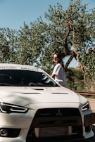 A person wearing sunglasses stands beside a white Mitsubishi sports car. The car is parked near trees with lush foliage under a clear blue sky.
