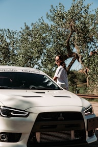 A person wearing sunglasses stands beside a white Mitsubishi sports car. The car is parked near trees with lush foliage under a clear blue sky.