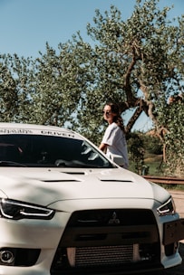 A person wearing sunglasses stands beside a white Mitsubishi sports car. The car is parked near trees with lush foliage under a clear blue sky.