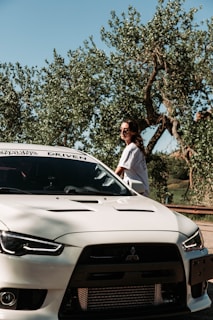 A person wearing sunglasses stands beside a white Mitsubishi sports car. The car is parked near trees with lush foliage under a clear blue sky.