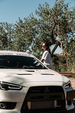 A person wearing sunglasses stands beside a white Mitsubishi sports car. The car is parked near trees with lush foliage under a clear blue sky.