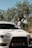 A person wearing sunglasses stands beside a white Mitsubishi sports car. The car is parked near trees with lush foliage under a clear blue sky.