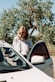 A happy customer standing next to a premium used car in a sunny outdoor setting.