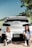 Two young women are sitting on the ground in front of a white car with a visible Colorado license plate. They are casually dressed in white shirts and jeans, with a tree and greenery in the background under a clear blue sky.