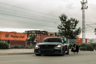 A sleek black car is parked on the side of a road, next to a young person squatting. Behind them, several large shipping containers, including one marked 'Hapag-Lloyd', are stacked against a backdrop of industrial looking buildings. Overhead, power lines are visible, and the scene is dominated by an overcast sky. A tall utility pole and a tree are also present near the road.