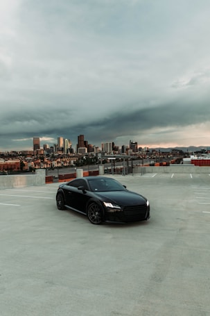 A sleek black car is parked in an open parking lot on a cloudy day with an urban skyline in the background. The scene is spacious and calm, with a large expanse of concrete surrounding the vehicle. The city buildings are illuminated by the soft light of sunset, creating a contrast between the industrial foreground and natural sky.