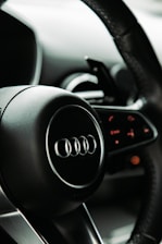 A close-up view of a car steering wheel featuring the Audi logo, with intricate details on the dashboard controls in the background. The design focuses on sleek, modern aesthetics with smooth black surfaces.