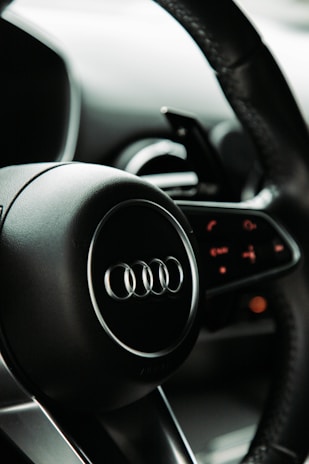 A close-up view of a car steering wheel featuring the Audi logo, with intricate details on the dashboard controls in the background. The design focuses on sleek, modern aesthetics with smooth black surfaces.