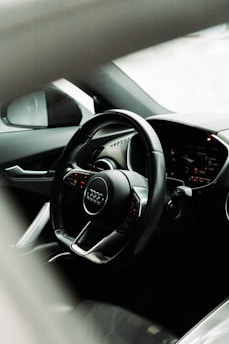 A car interior showcasing a sleek design with a focus on the steering wheel featuring the Audi logo. The dashboard displays illuminated indicators and controls, suggesting a modern vehicle with advanced technology.